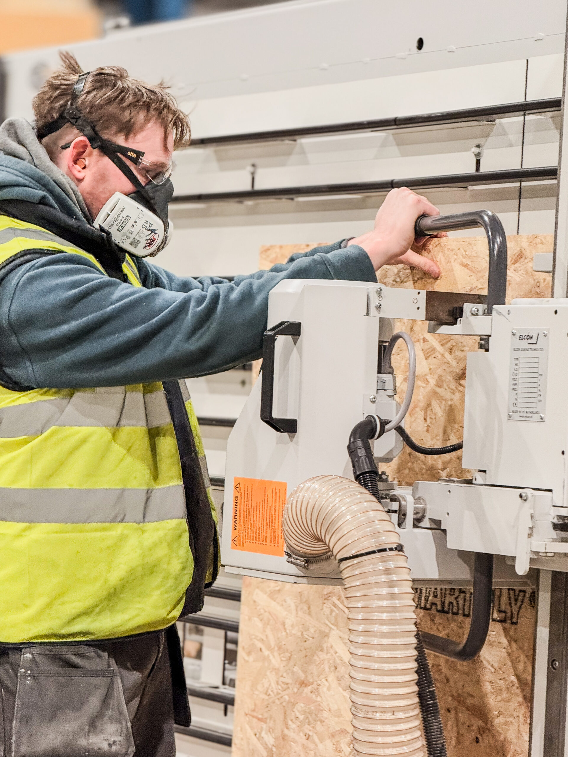 Man operates vertical panel saw, wearing dust mask and high-visibility yellow jacket.