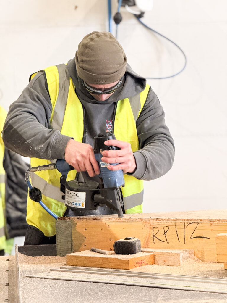 A man with a yellow high-visibility jacket uses a nail gun to assemble a timber frame panel.
