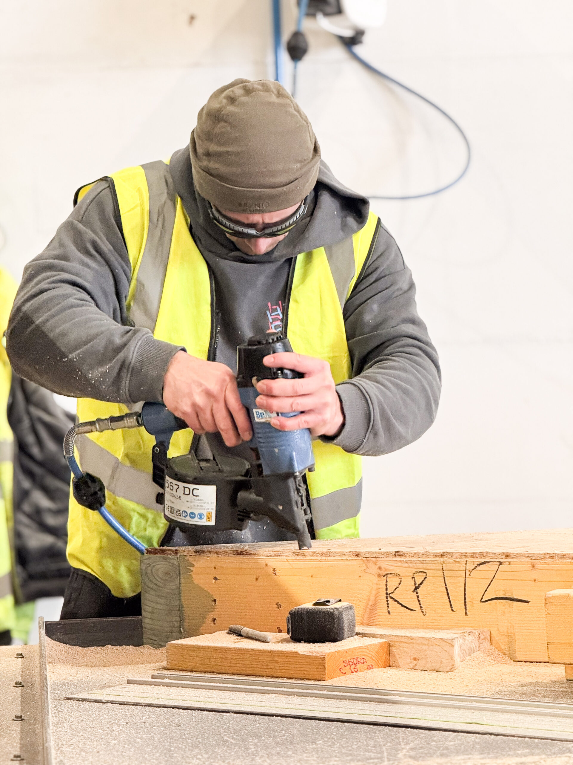 A man with a yellow high-visibility jacket uses a nail gun to assemble a timber frame panel.