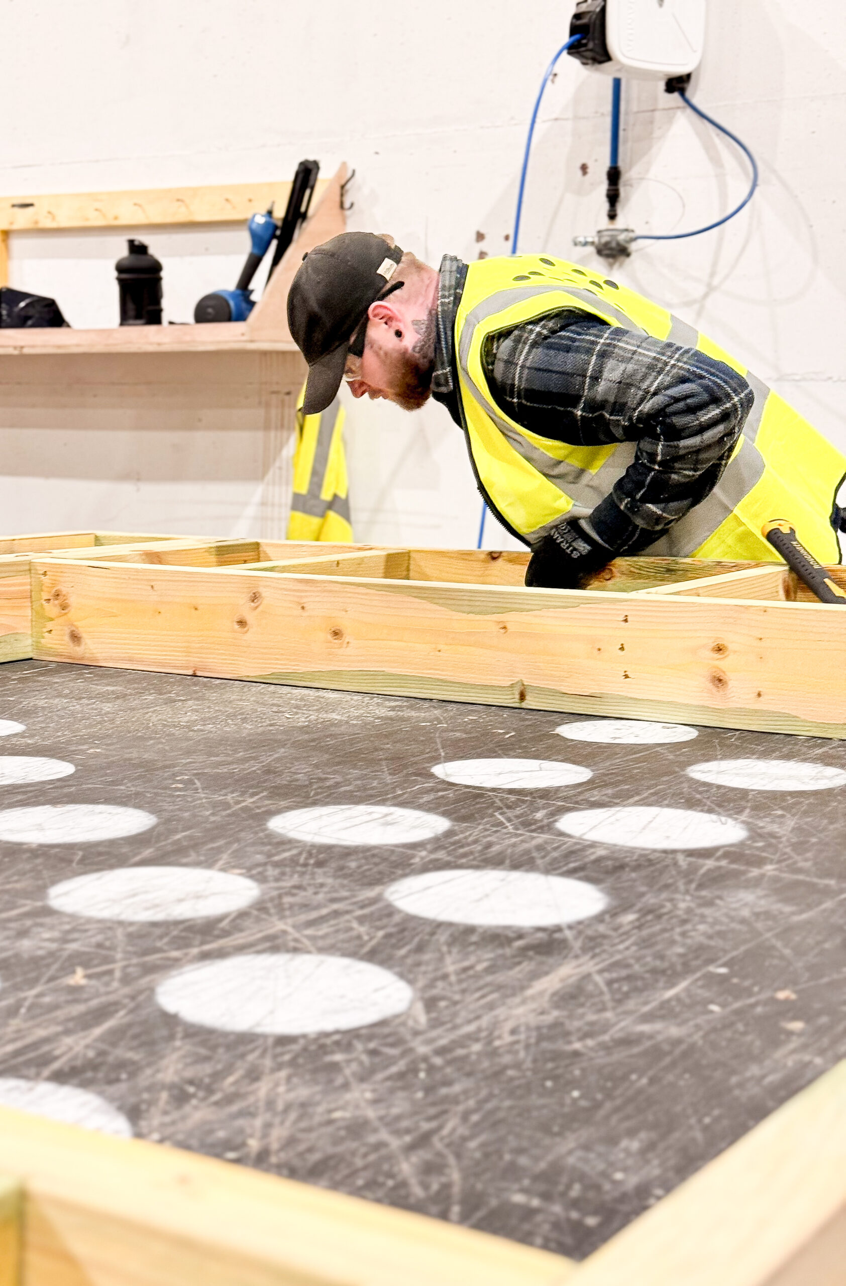 A man in a yellow high-visibility jacket and black baseball cap inspects a timber frame structure on a work table.