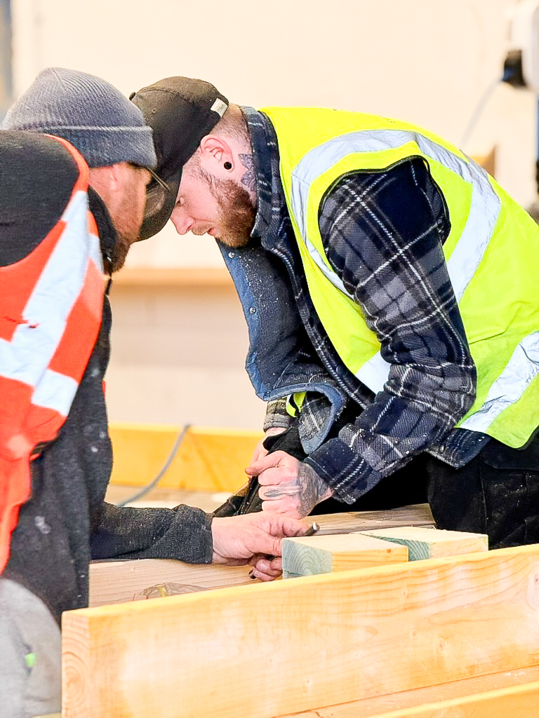 A man in a yellow high-visibility jacket and black baseball cap uses his fist to knock down an element of timber structure on a work table.