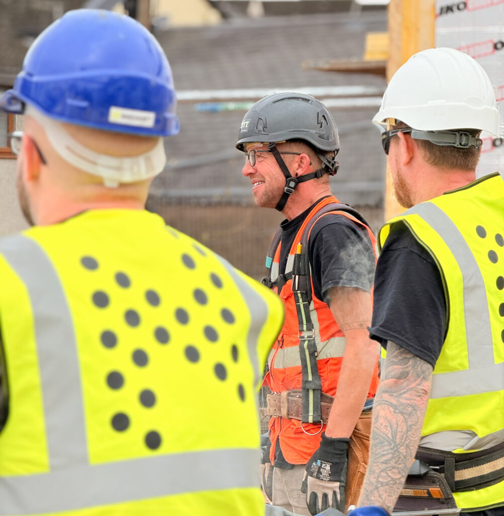 Three men wearing high-visibility vests, and hard hats, one of the men is wearing a tool harness with an assortment of tools for timber frame erection.