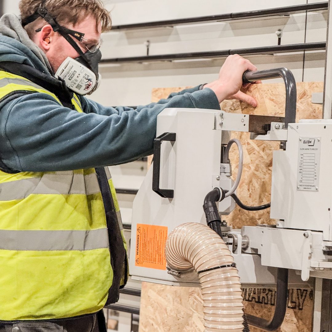 Man operates vertical panel saw, wearing dust mask and high-visibility yellow jacket.